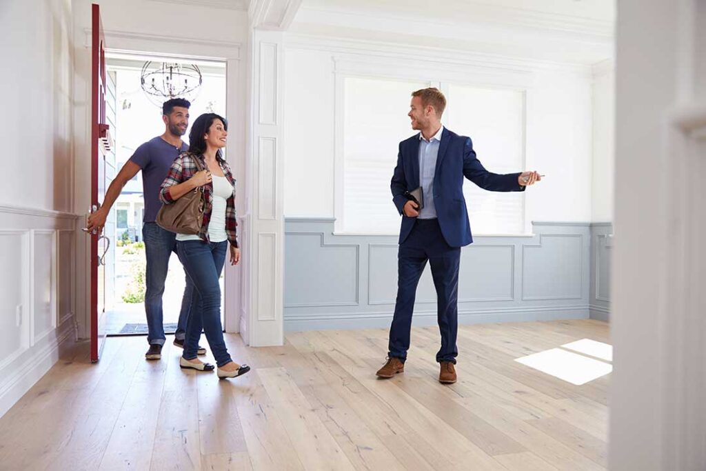 A real estate agent and couple review property documents at a kitchen table beside a home inspection report.